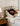 A woman with curly dark hair rests asleep on a floral-patterned bed with a gold metal headboard, next to a wooden bedside table with a lamp and glasses. Her Bon Terre Cotton Blossom & Lavender Room & Body Mist bottle is also on the table.